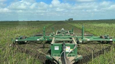 Una grada agrícola verde, tirada por un tractor, atraviesa un campo de cultivos verdes bajo un cielo parcialmente nublado.