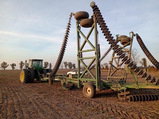 Un tractor verde arrastra una gran grada de discos desplegada, con secciones laterales elevadas, en un campo al anochecer.
