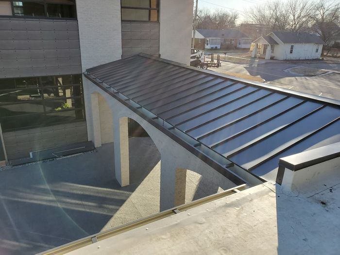 Apartment walkway with a sloped metal roof and concrete support columns beside a building.