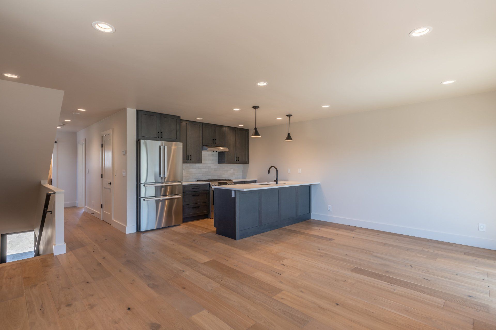 An empty living room with hardwood floors and a kitchen with stainless steel appliances.