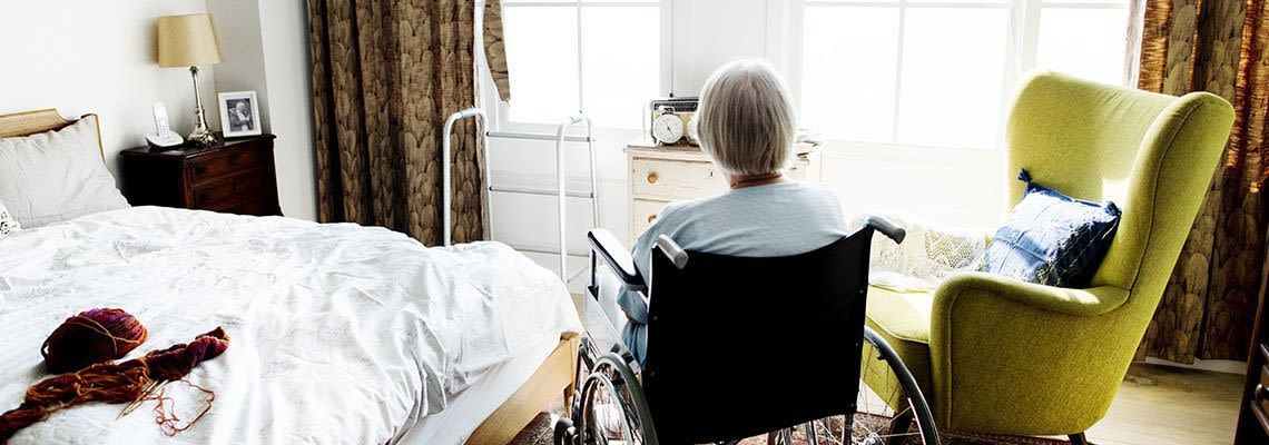 An elderly woman in a wheelchair is sitting in a bedroom next to a chair.