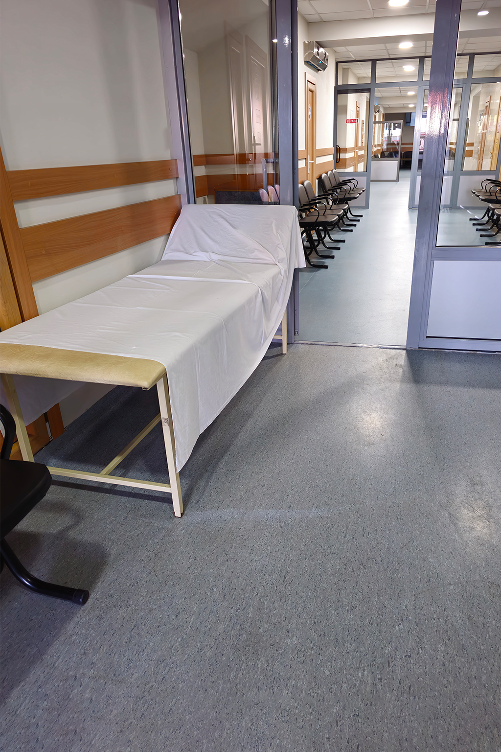 Examination table in a hospital hallway with chairs and glass doors.