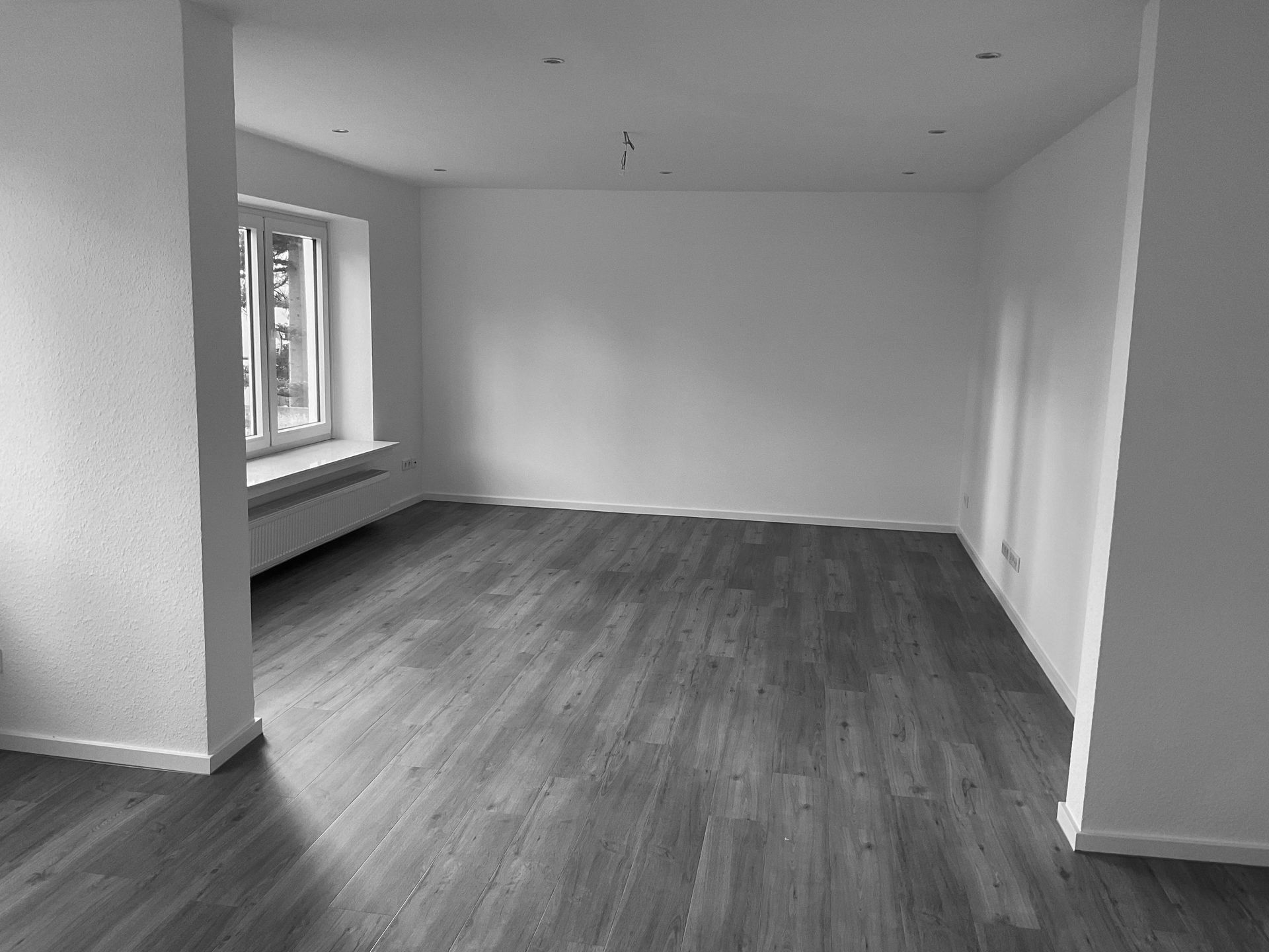 Ein Schwarzweißfoto eines leeren Wohnzimmers mit Hartholzböden und weißen Wänden.
