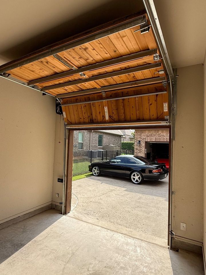 Open garage with raised wooden door, showing driveway and a black car parked outside.