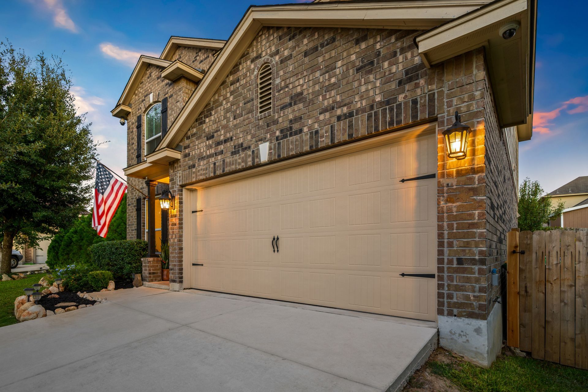Brick suburban house with double garage, front walkway, American flag, and lit porch at dusk