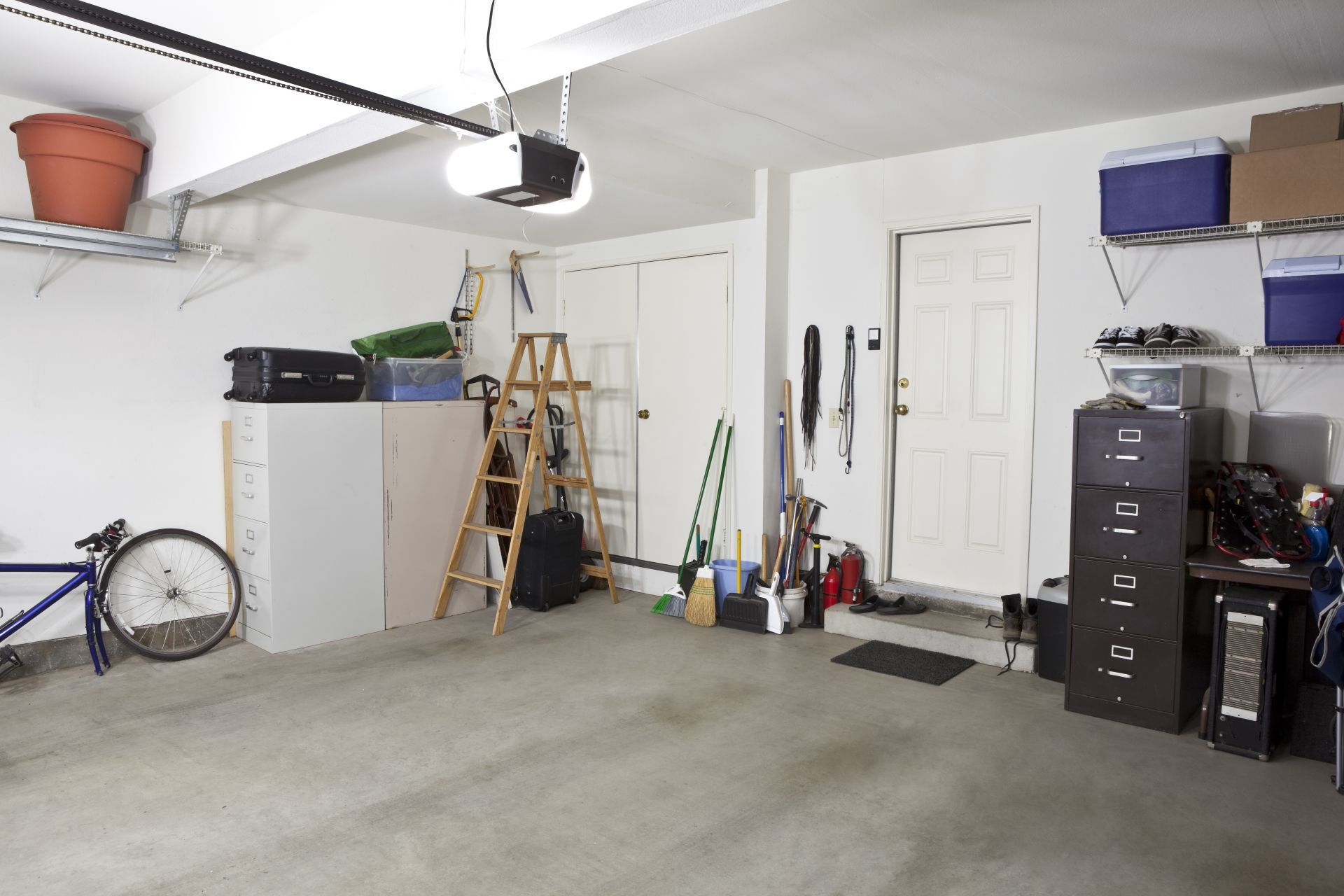 Garage interior with concrete floor, storage shelves, tools, bicycle wheel, and a closed white door