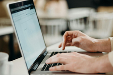Hands typing on a laptop keyboard; screen shows website in light colors, indoors.