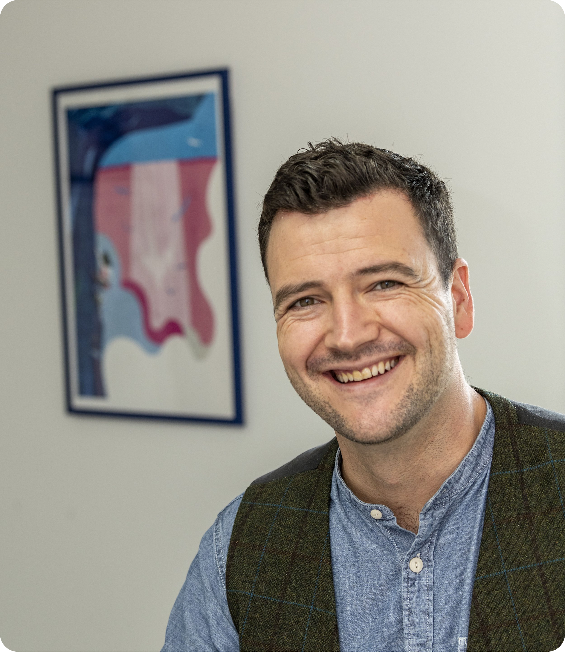 Man smiling, leaning forward, wearing a blue shirt and green vest, with colorful abstract art in the background.