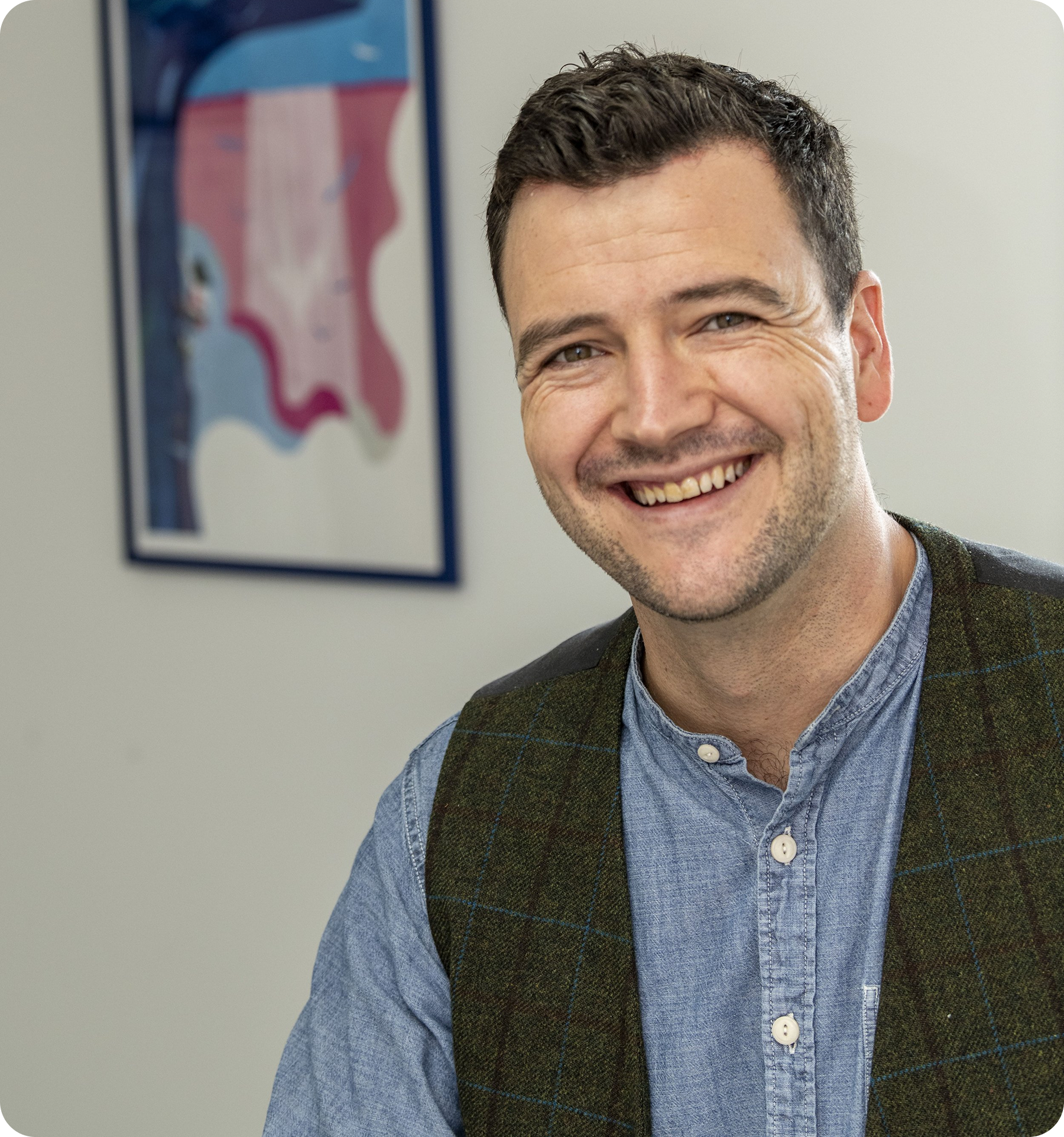 Man smiling, wearing a blue shirt and green vest, in front of a framed abstract artwork.