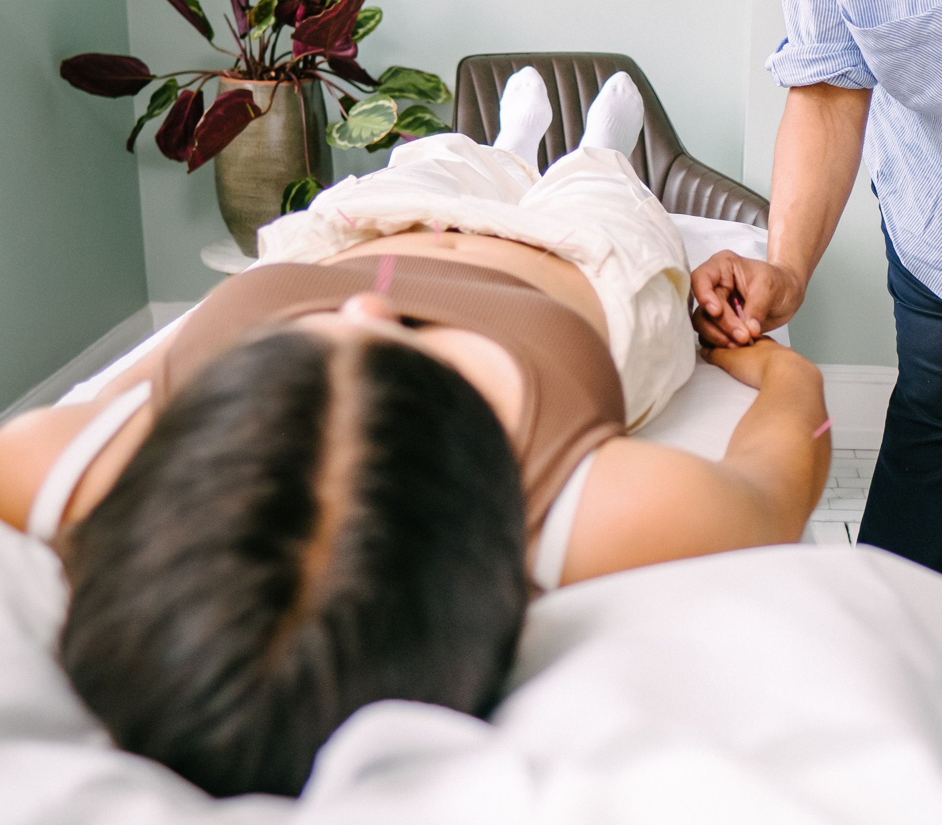 A female patient is lying on her back, with some needles in, as a practitioner to the right is adding a new point.