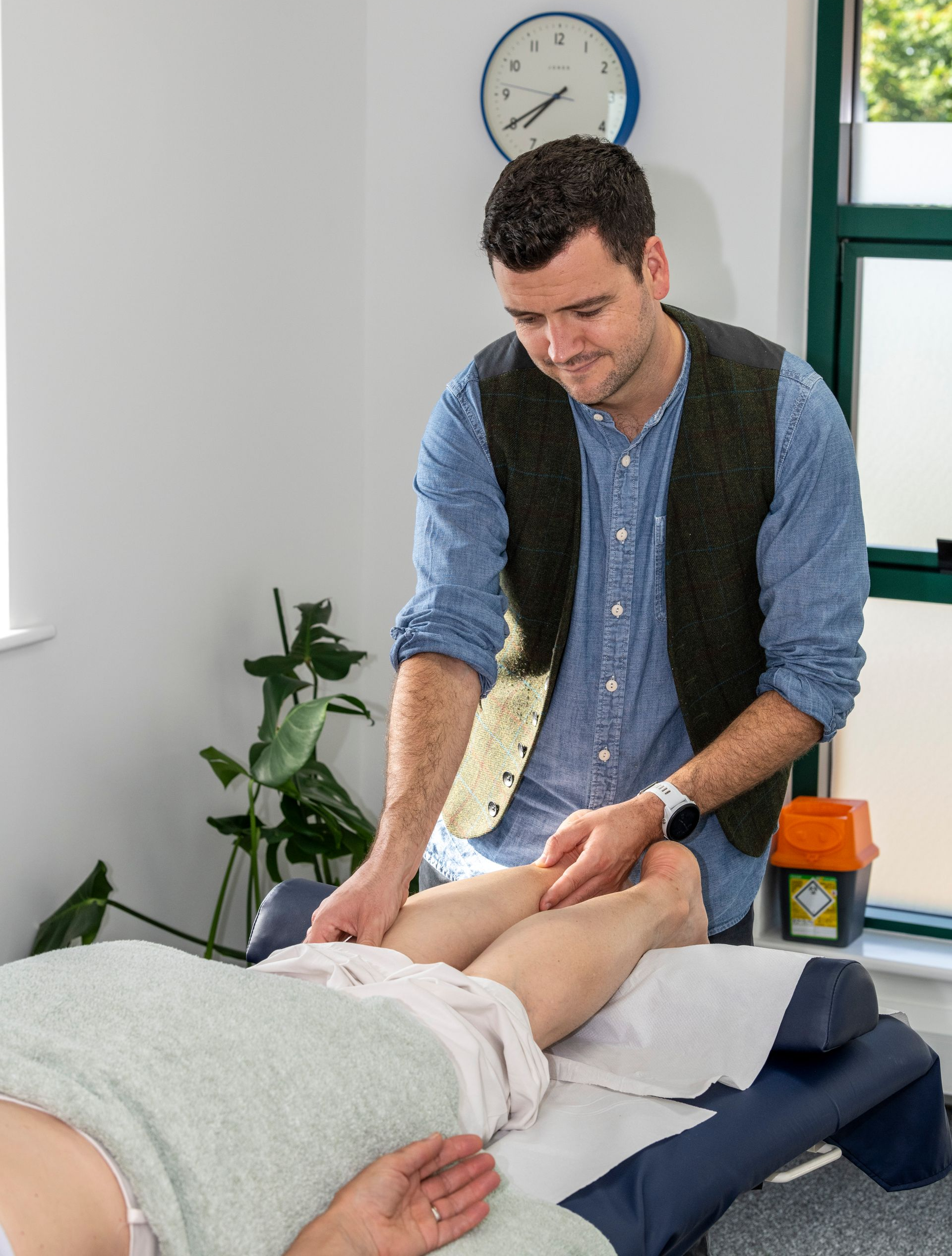 Man examining patient's leg on a treatment table. He wears a blue shirt and vest in a bright room with a clock and plants.
