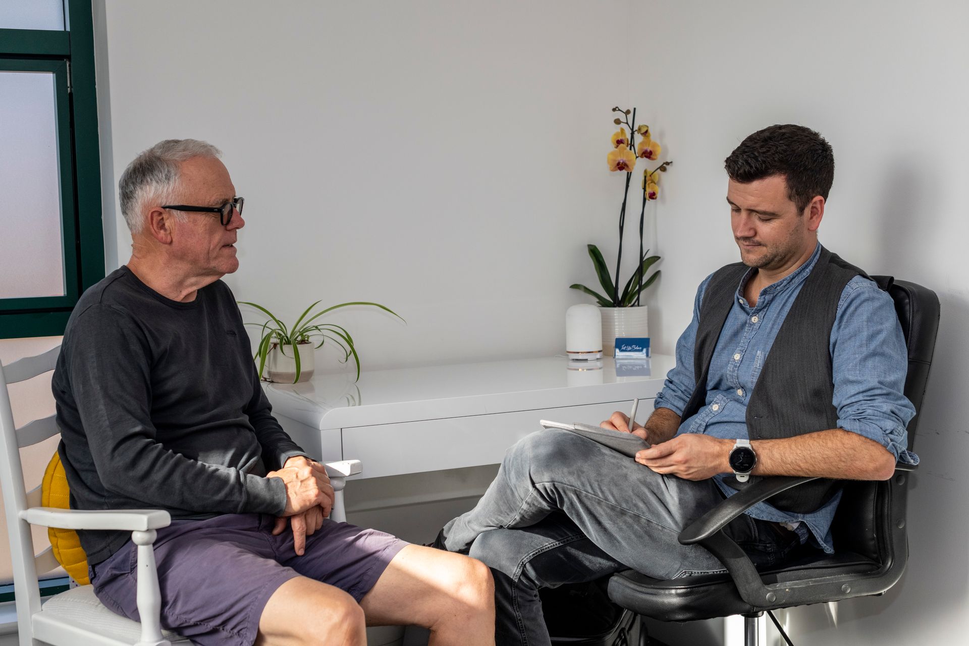 An acupuncture consultation where a male patient is sitting to the left of the acupuncturist whilst explaining his health.