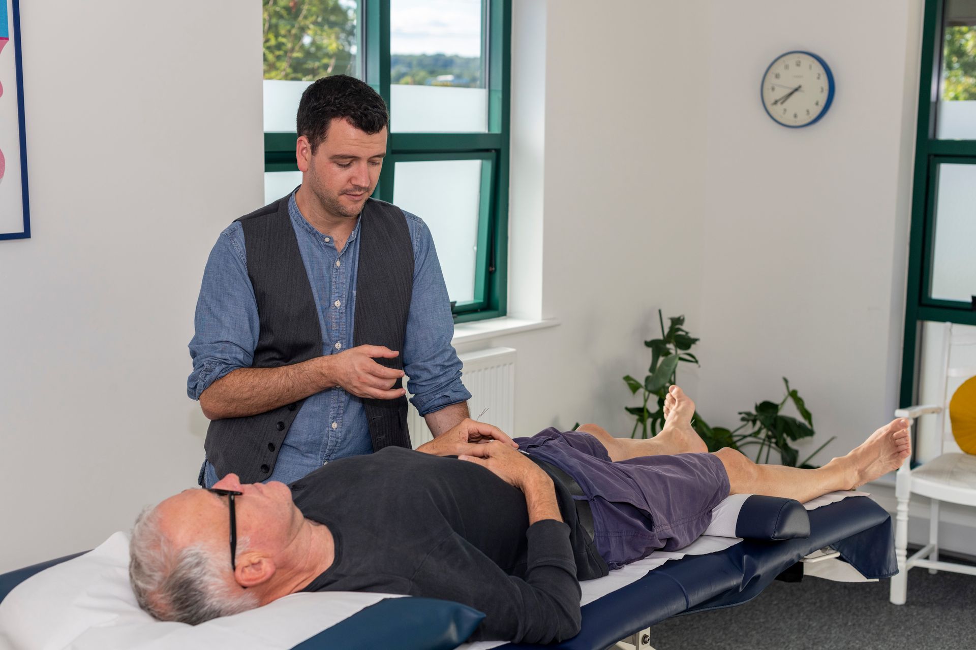 Man examining a person lying on a therapy bed in a light-filled room.