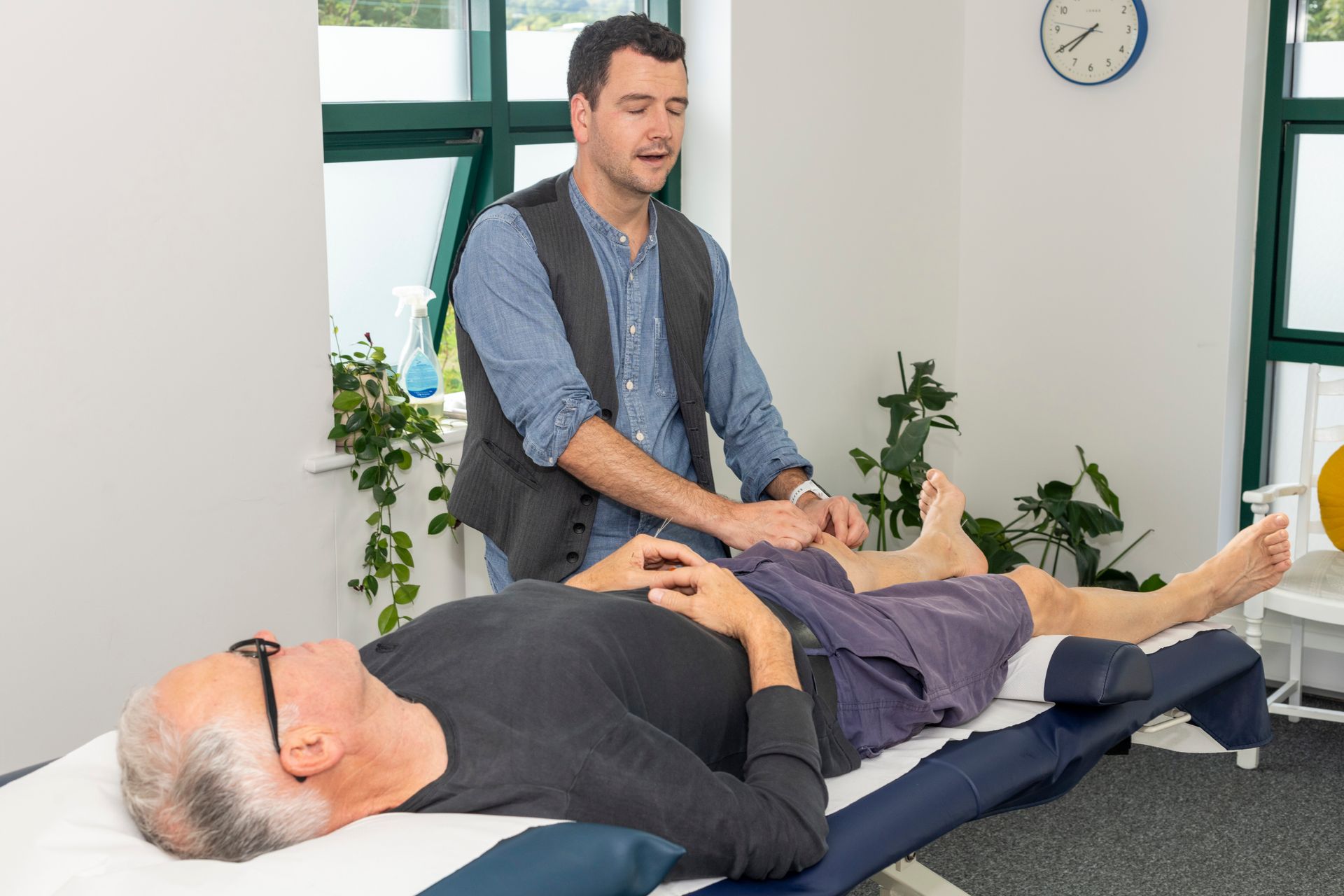 An acupuncturist, Josh, is treating a male patient who is lying down by massaging his knee.