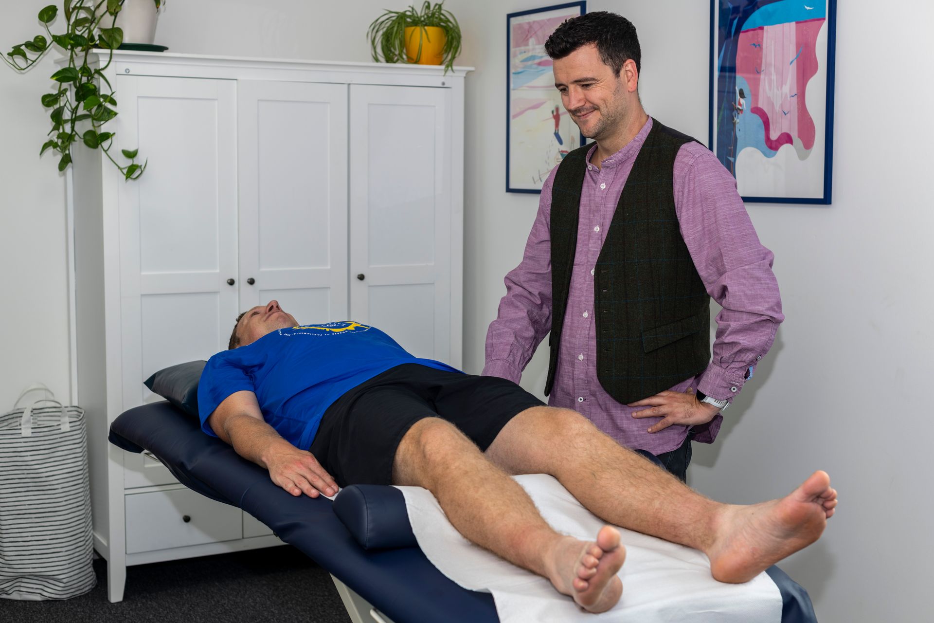 Man on therapy table, another man standing over him. Interior shot, white wall.