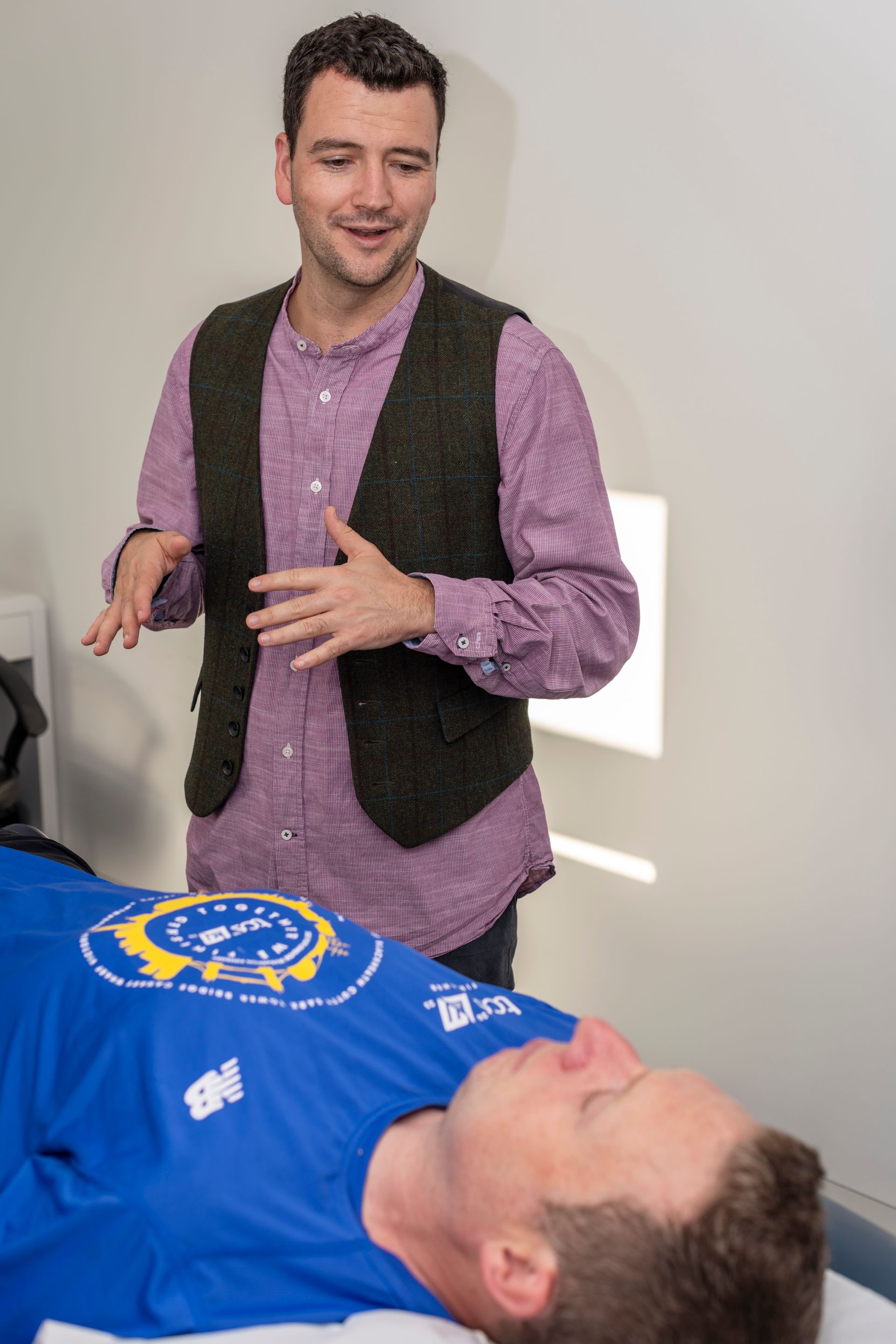 The acupuncturist, stood above a patient, is talking to the patient with his arms raised in an expressive and friendly way.