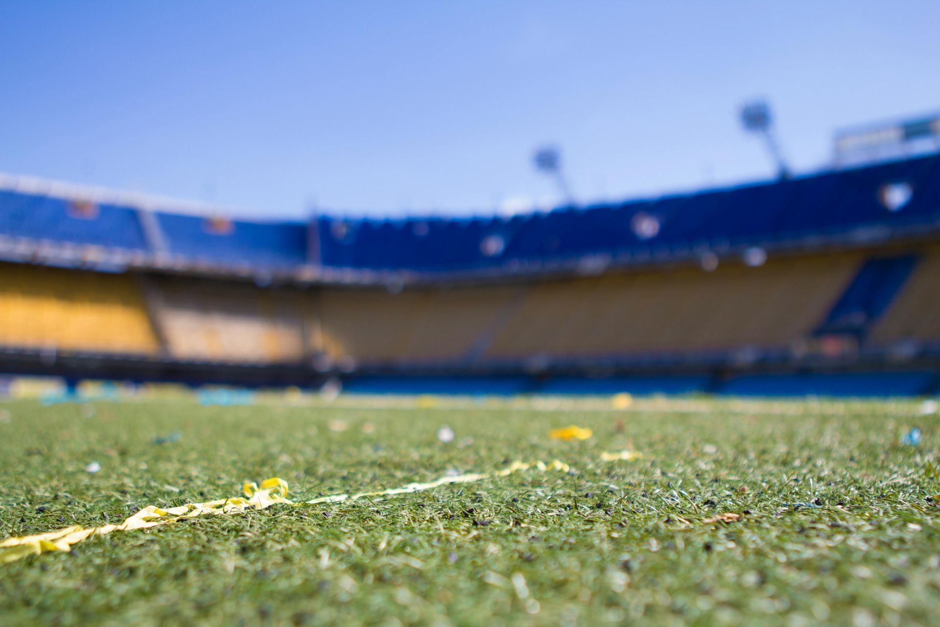 Green soccer field, yellow line, and blurred stadium with blue and yellow seating under blue sky.