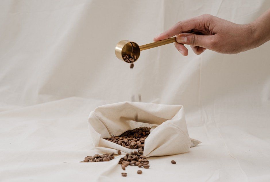 Hand pouring coffee beans from a golden scoop into an open bag on a cream-colored surface.