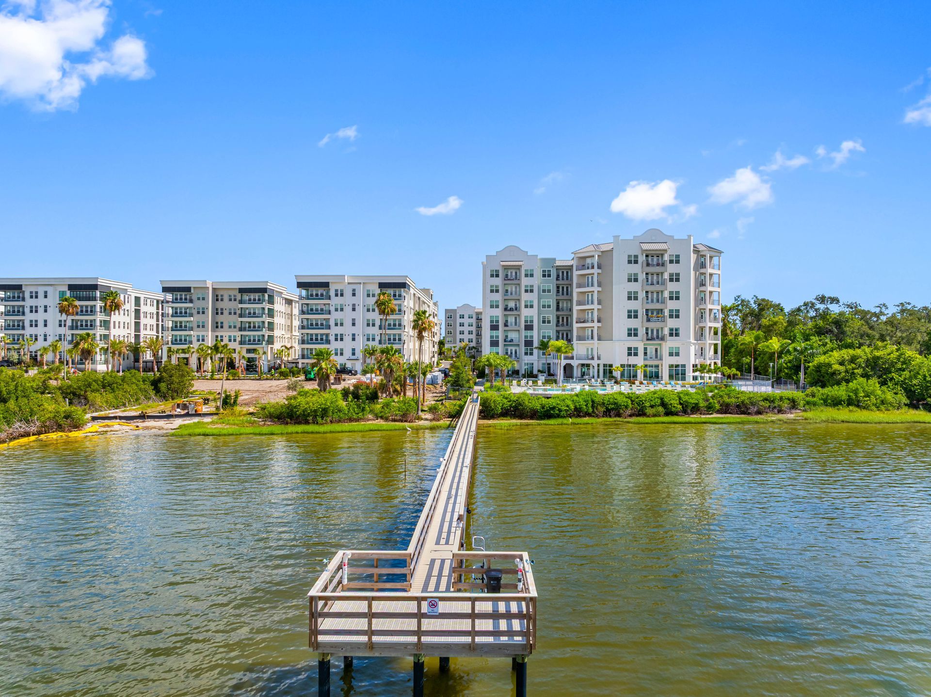 Pier extending into water, leading to waterfront condos under blue sky.
