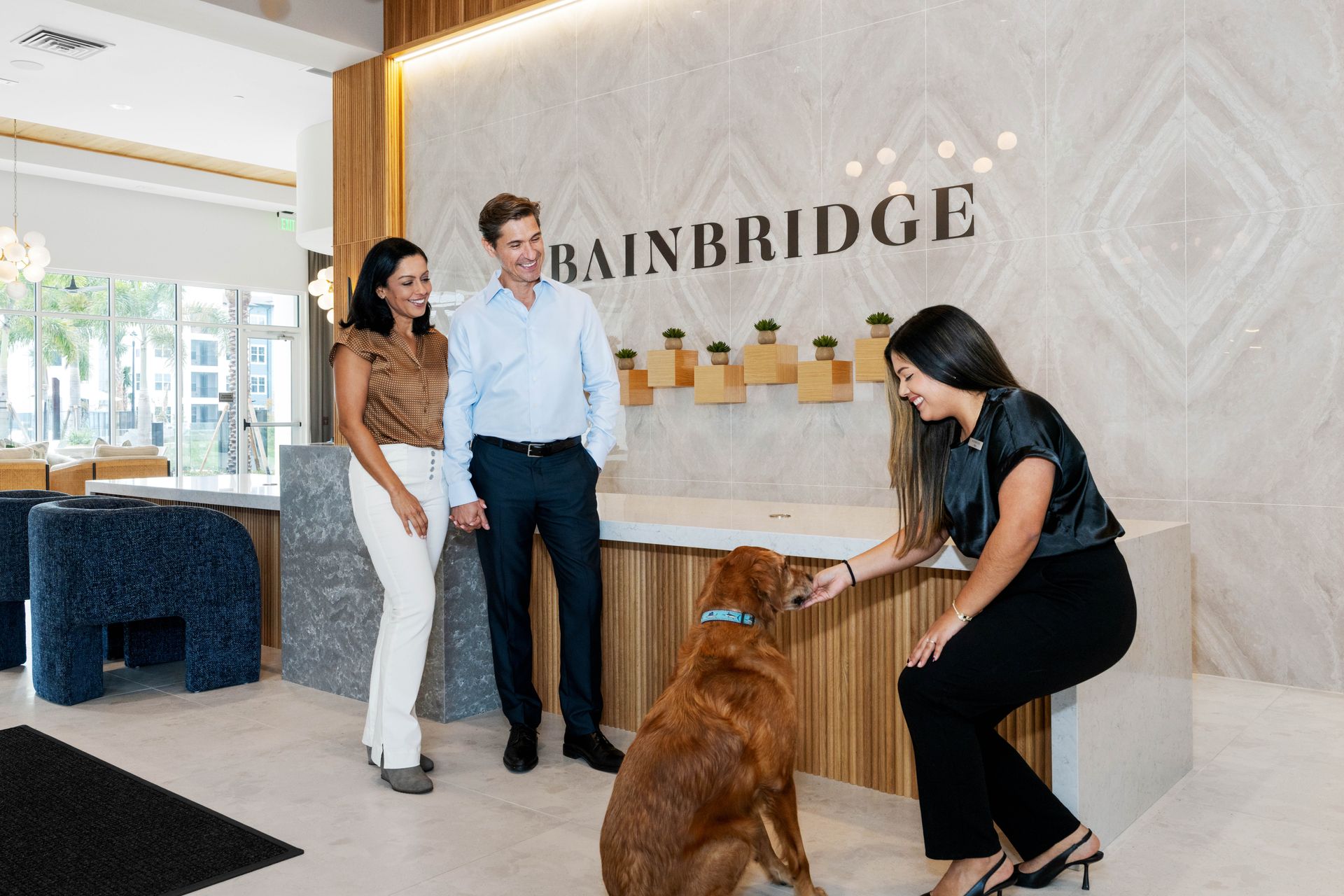People at a Bainbridge reception desk, petting a golden retriever. Light interior with logo.
