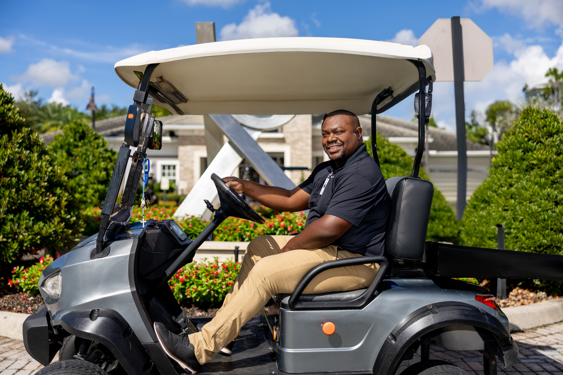 Man driving a gray golf cart, smiling, wearing a black shirt and khaki pants. Daytime.