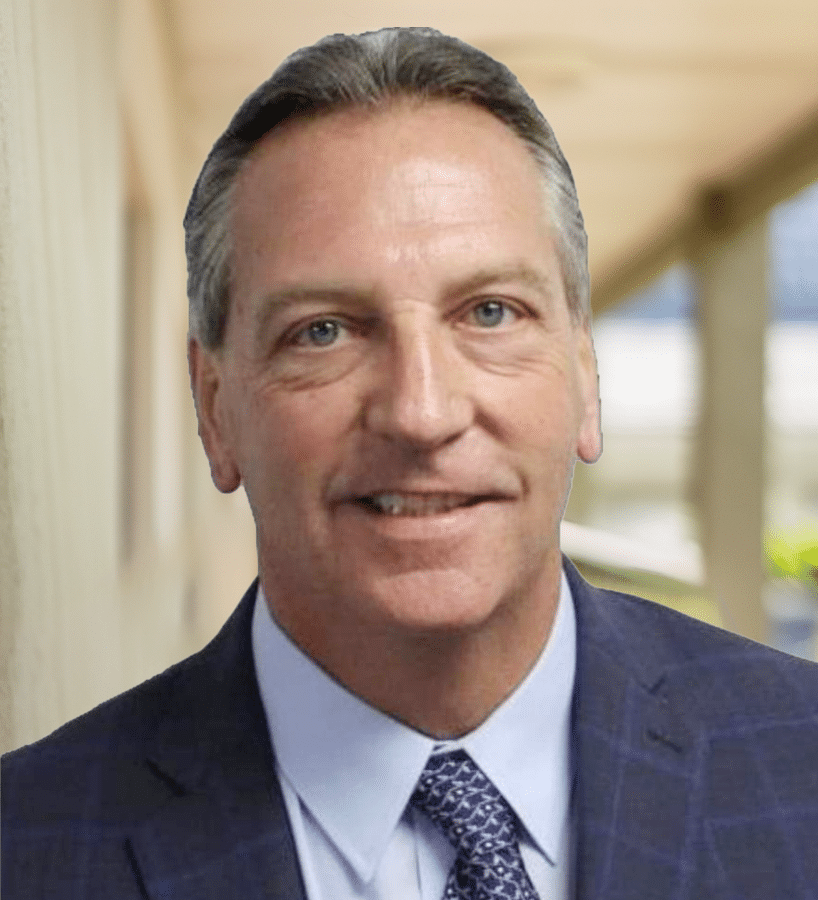 Man in a suit smiles, looking directly at the viewer. Blurred background.