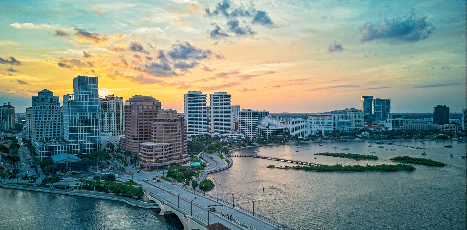 Sunset over a city skyline with water, bridge, and buildings.