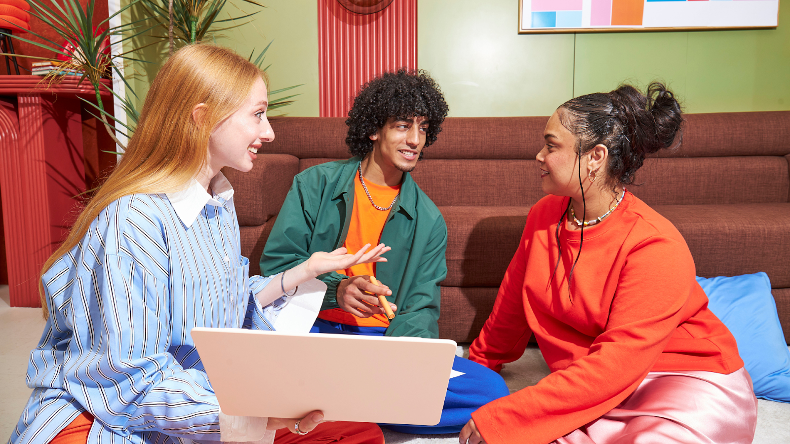 Three friends sit together on the floor in a room, discussing a laptop held by the person on the left.