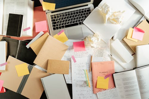 Overhead view of a cluttered desk with laptop, notebooks, papers, and sticky notes spread around