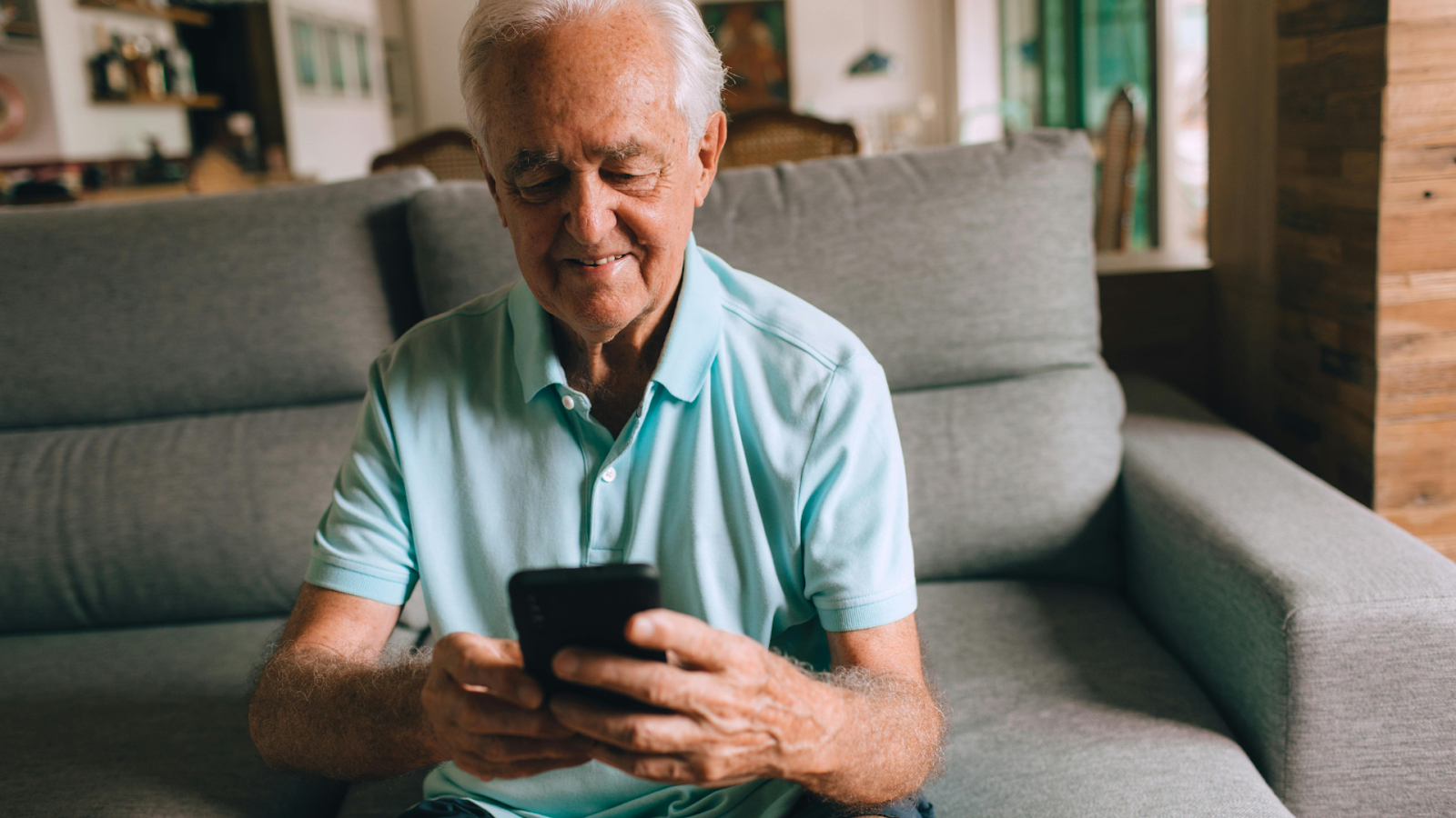An older person in a light blue polo shirt sits on a grey sofa, smiling while looking at a smartphone held in their hands.