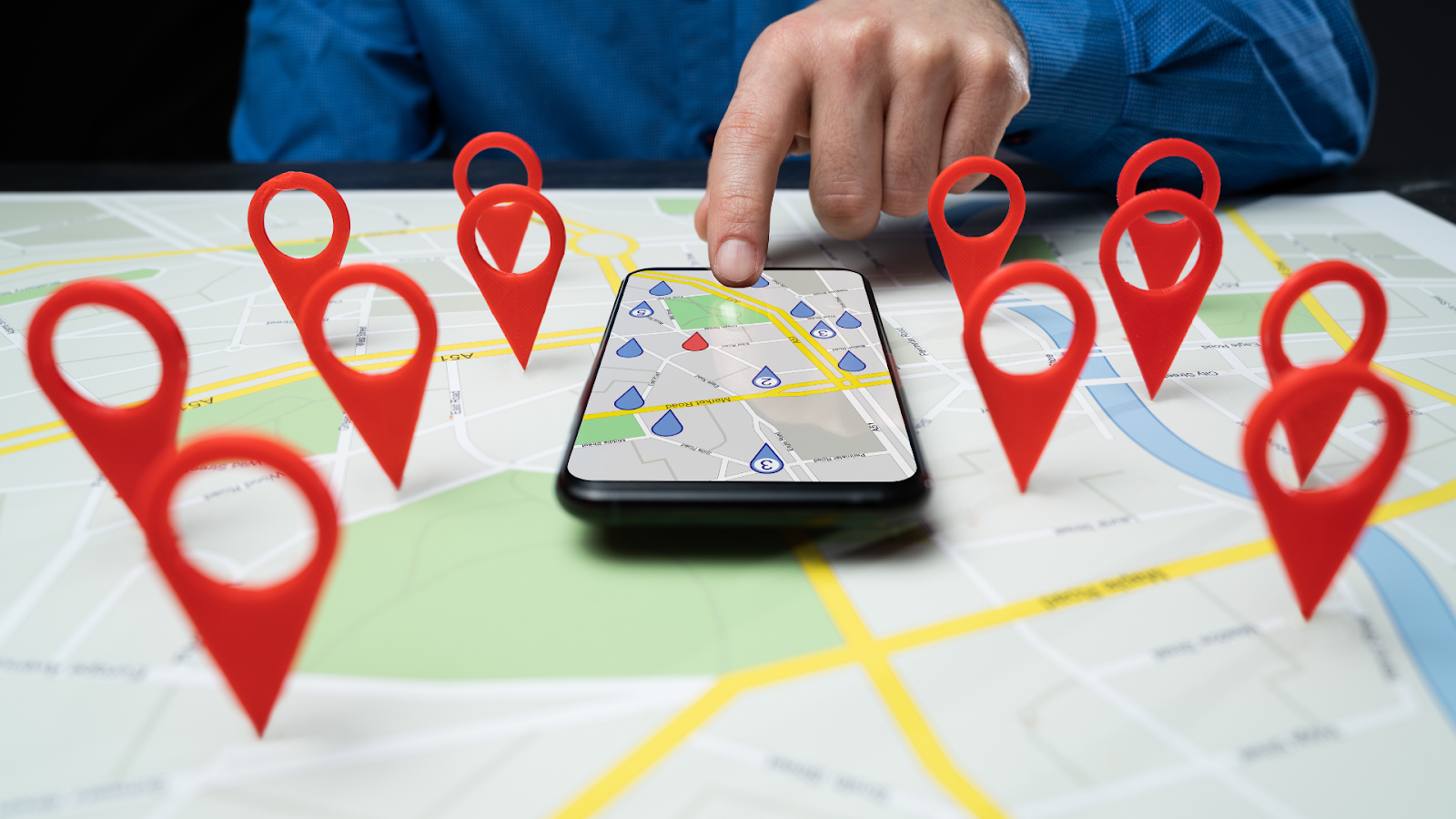 A hand touches a smartphone screen displaying a map, surrounded by several red 3D location pin icons on a paper map.