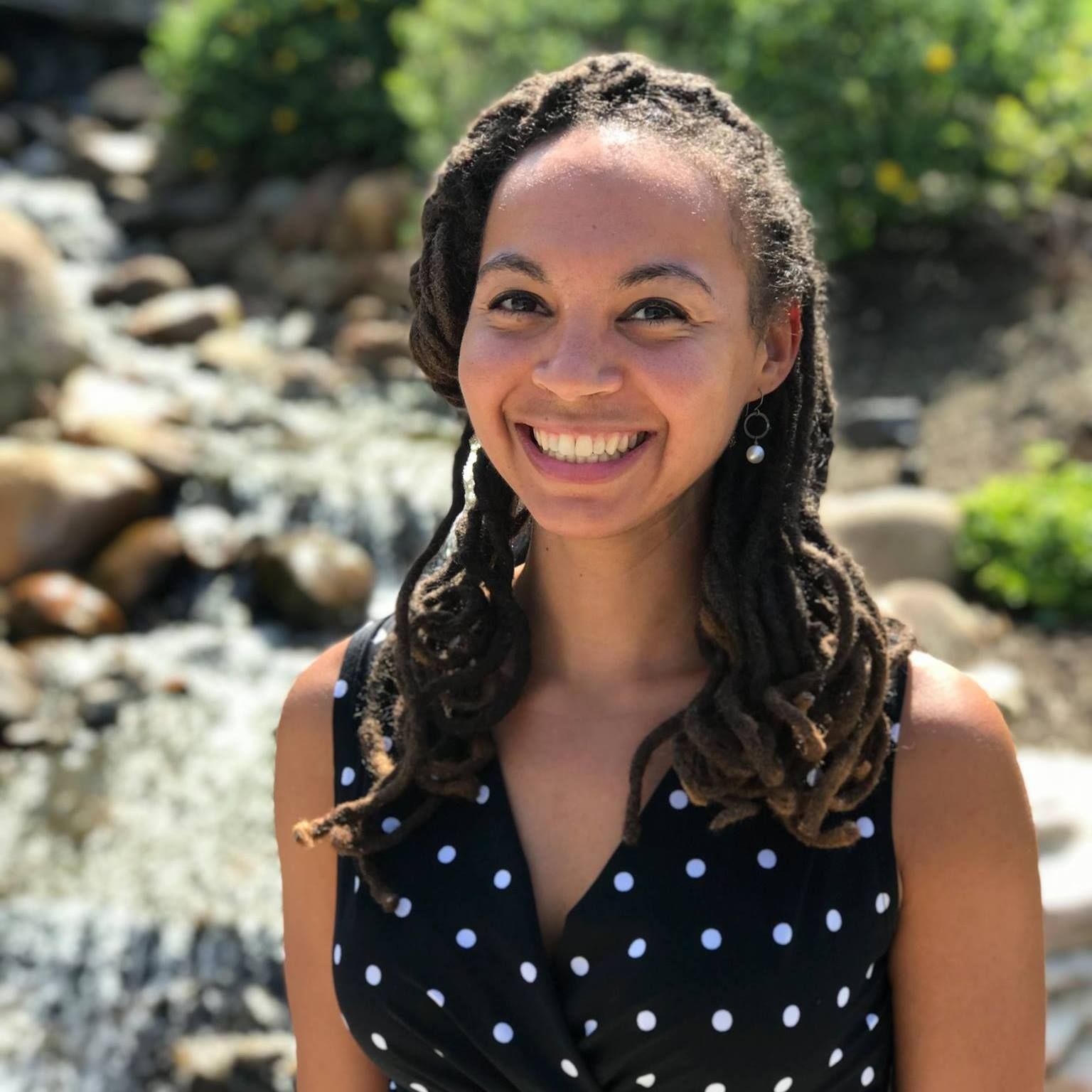 A woman in a black and white polka dot dress is smiling in front of a waterfall.