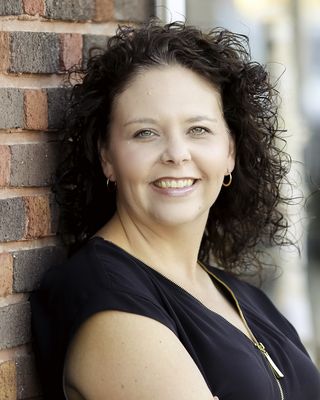 A woman with curly hair is leaning against a brick wall and smiling.