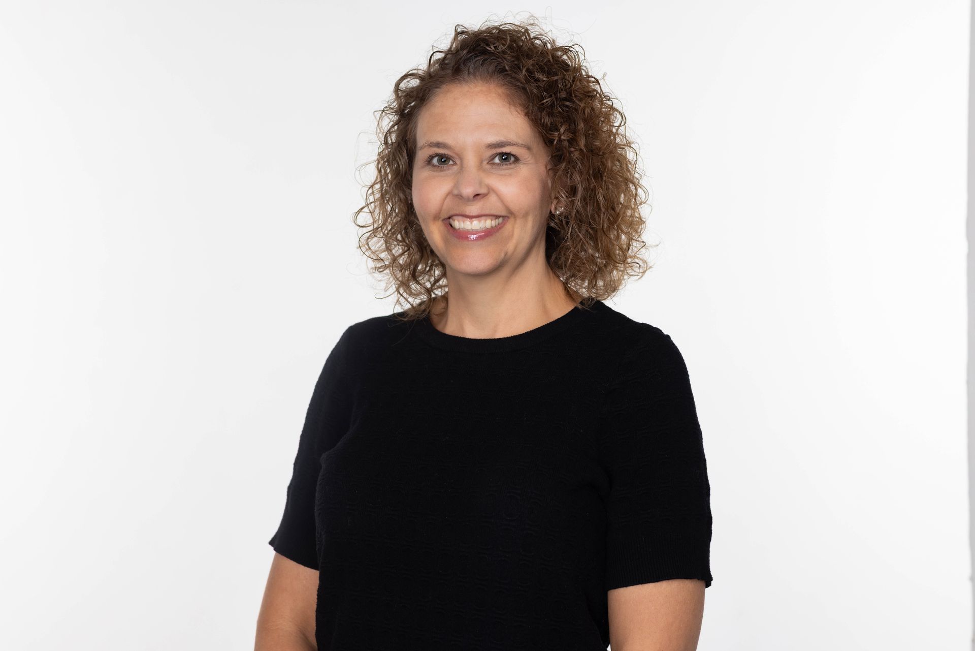 A woman with curly hair is leaning against a brick wall and smiling.