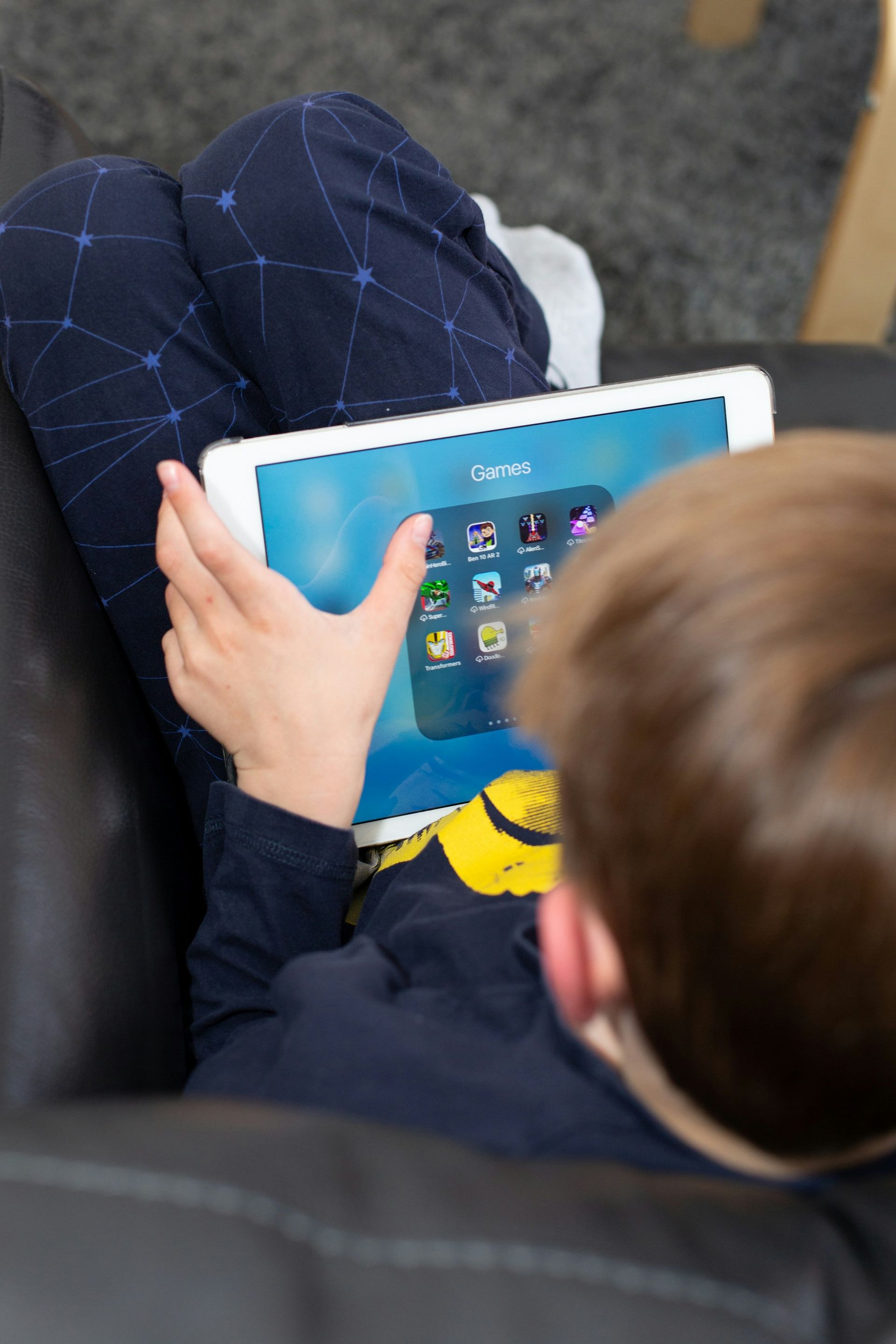 Child sitting, using a tablet, on a black couch. Blue clothing, light skin, various app icons on the screen.