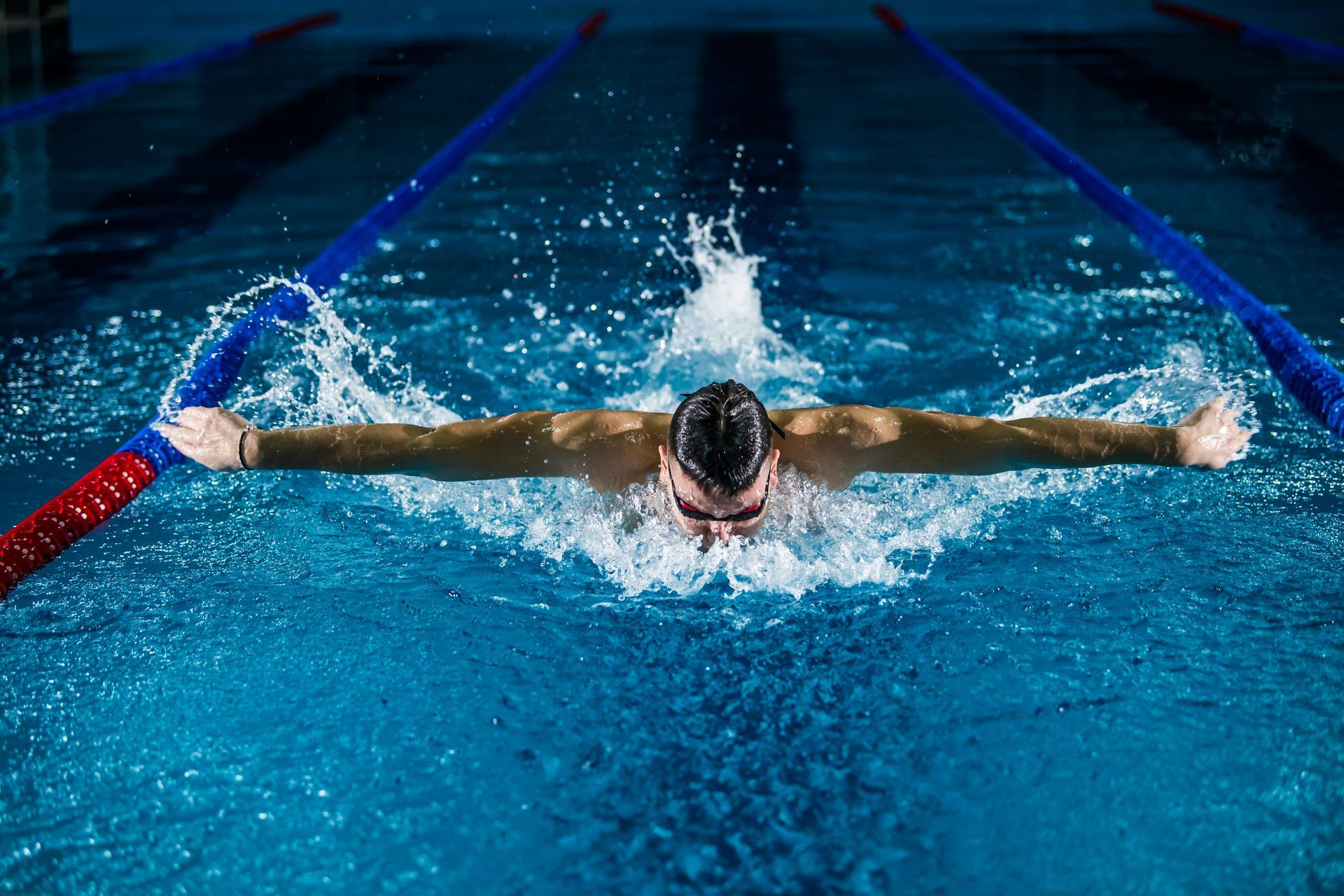 Swimmer in butterfly stroke in blue pool, arms outstretched, water splashing.