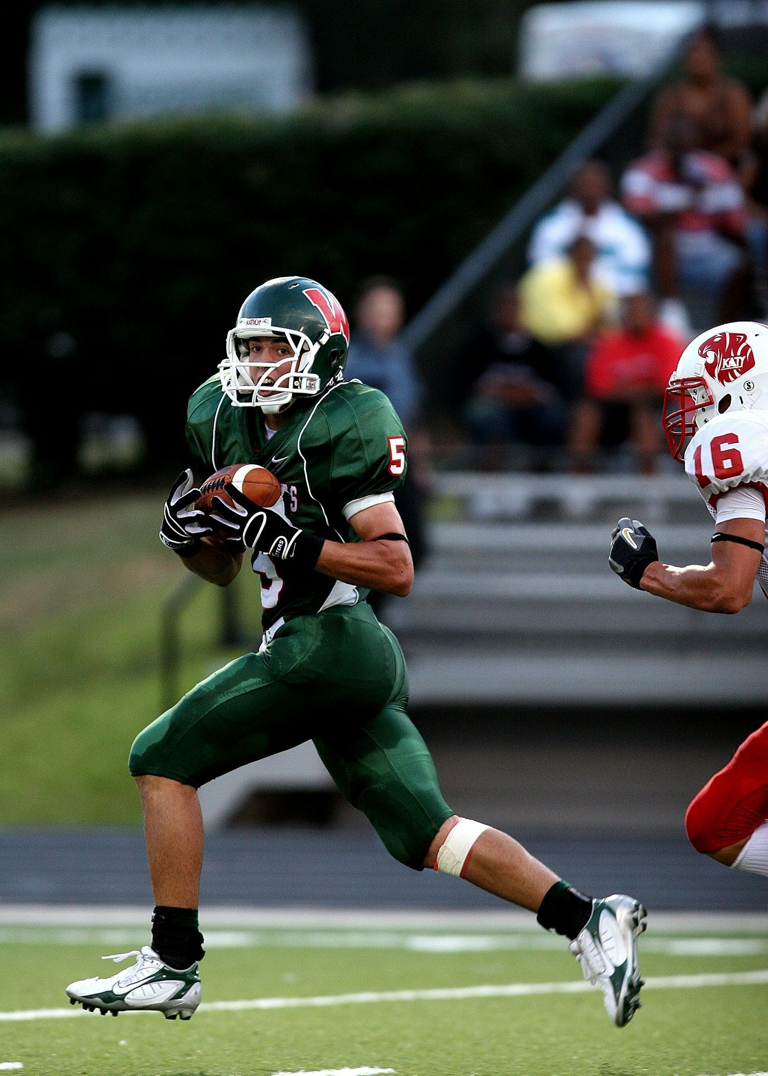 Football player in green uniform running with the ball, other player in red close behind.