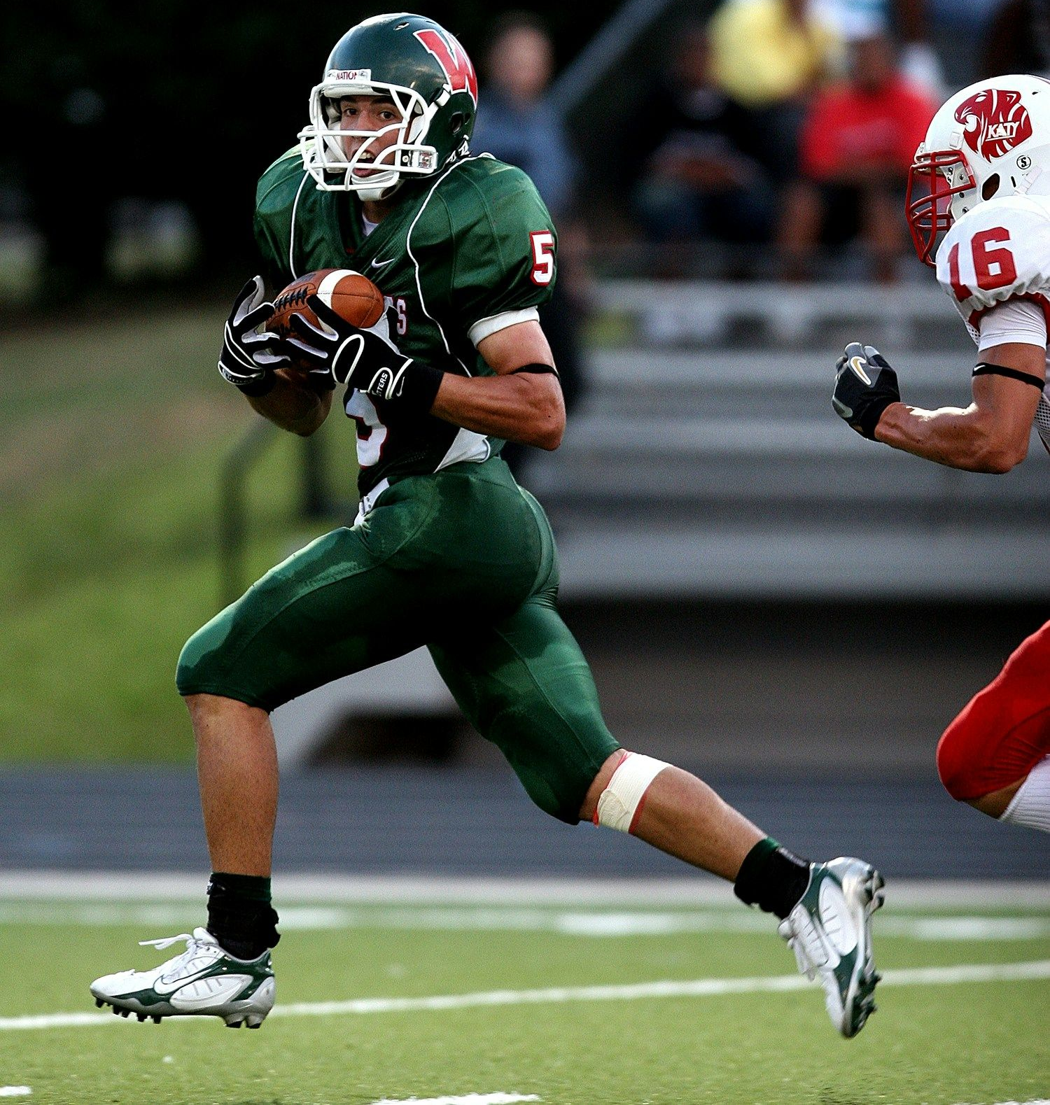 Football player in green uniform runs with the ball. An opposing player in red tries to tackle.
