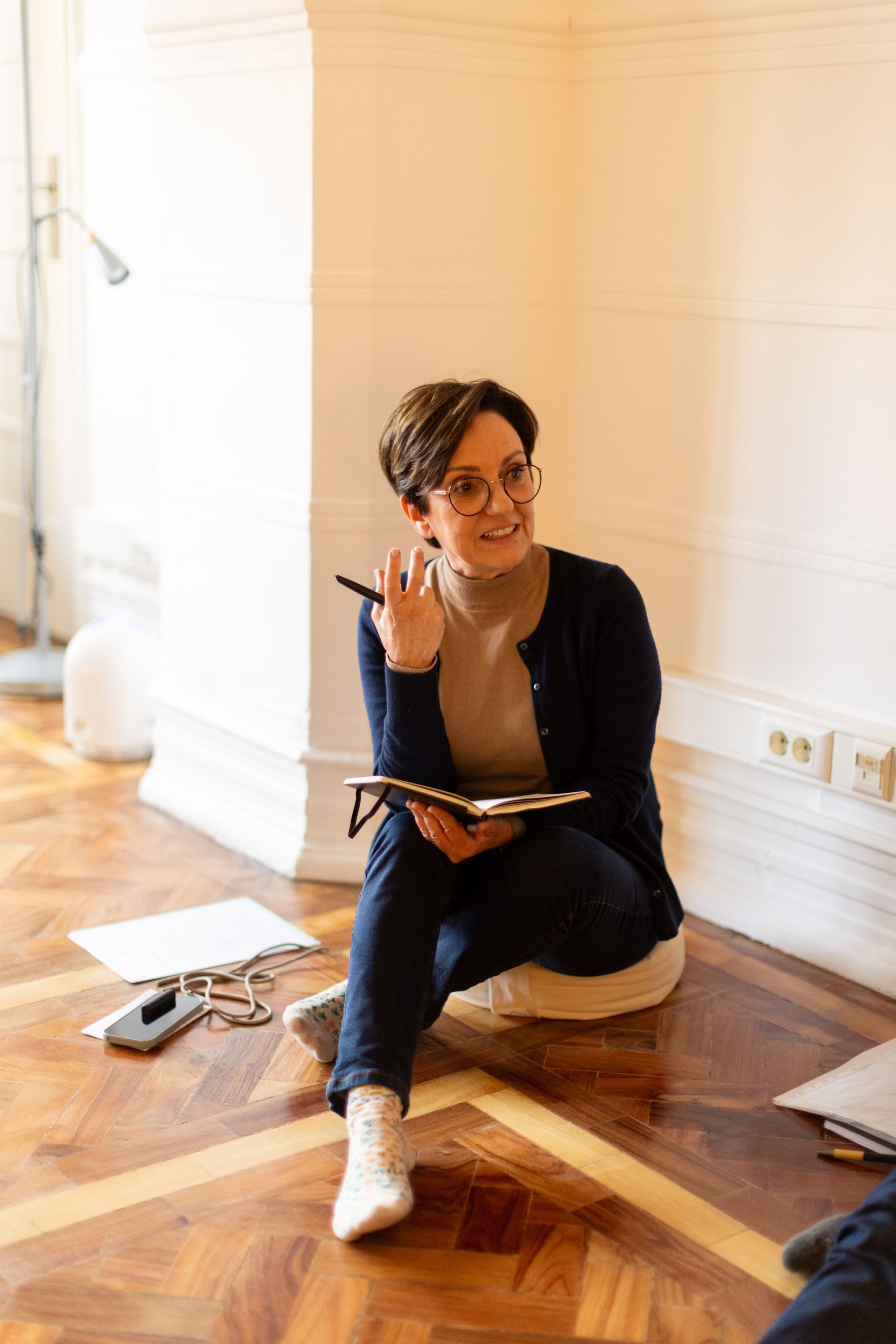 Trainer sitting on the floor with a notebook in hand, speaking and gesturing during a training session. The setting is informal and welcoming, with natural light and wooden flooring.