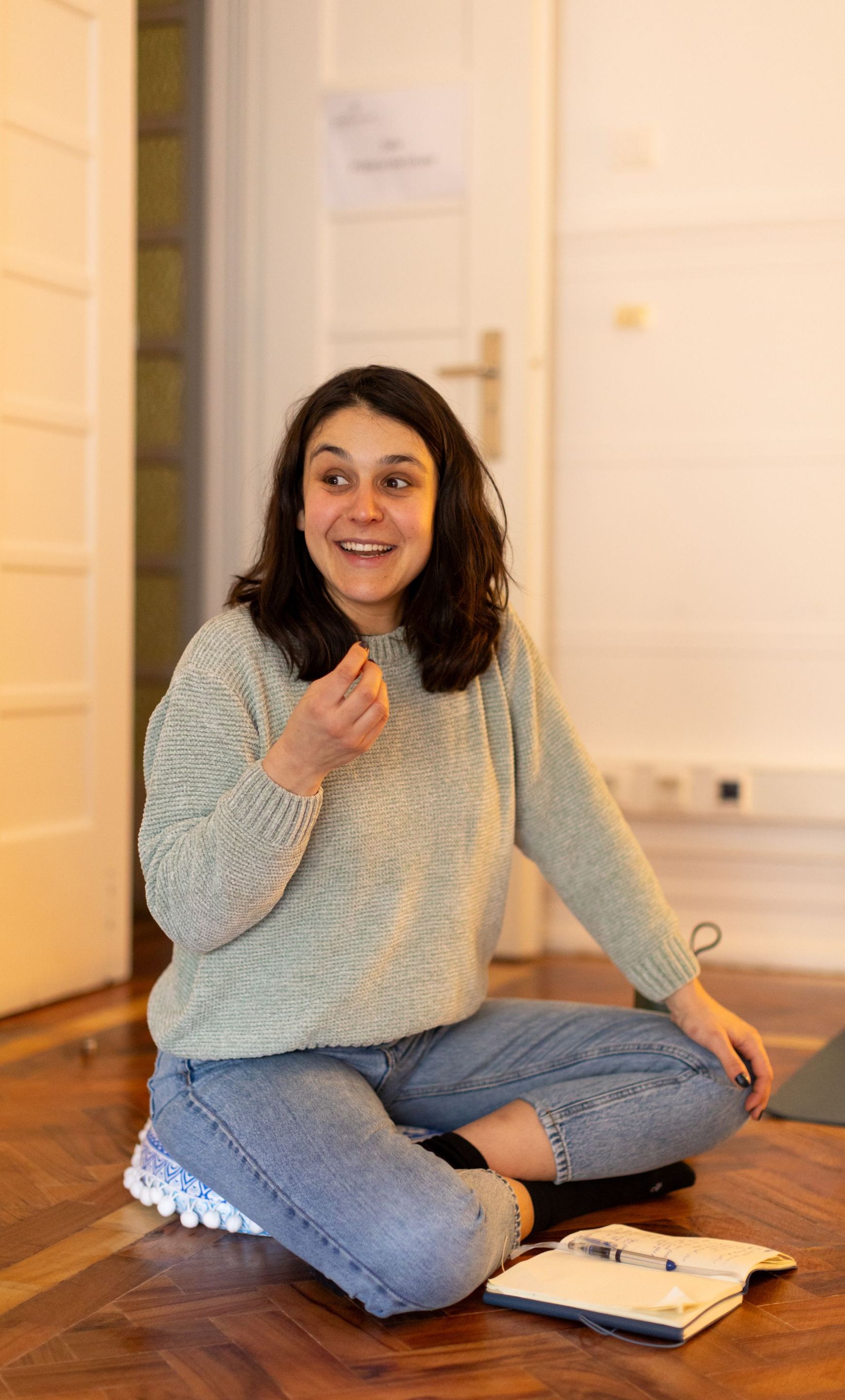 Trainer during the 'Voice and Body Language' module, sitting on the floor with a notebook in front of her, smiling and communicating with the group in a welcoming training space.
