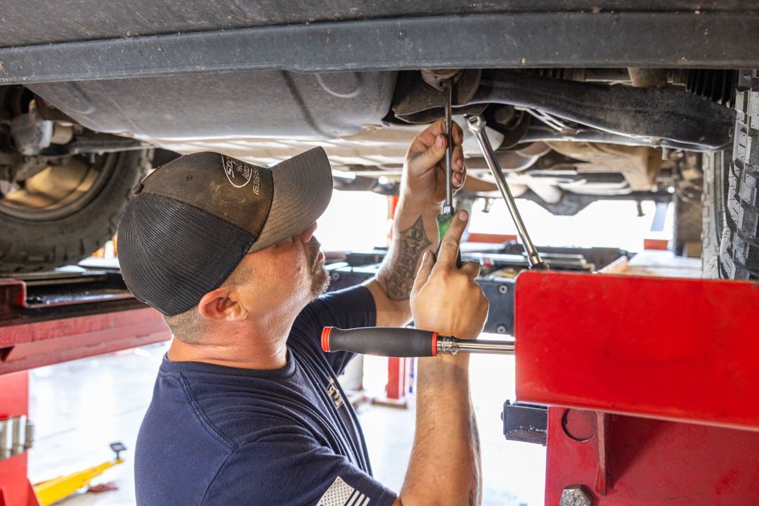 Mechanic under a car, working with tools. He wears a baseball cap and has tattoos on his arm.