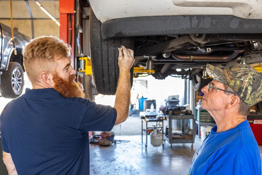 Mechanic showing a car's underside to a person in a garage, pointing at a tire.