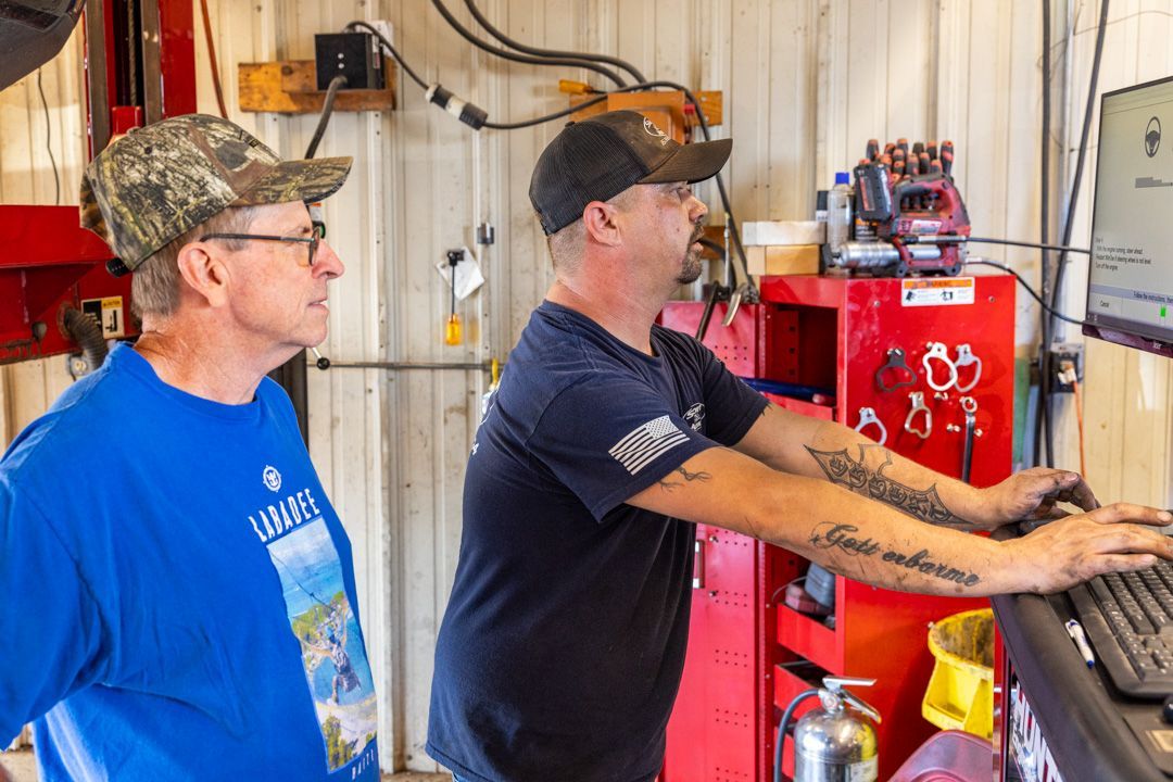 Two men in a garage looking at a computer. One types on a keyboard, the other looks on.
