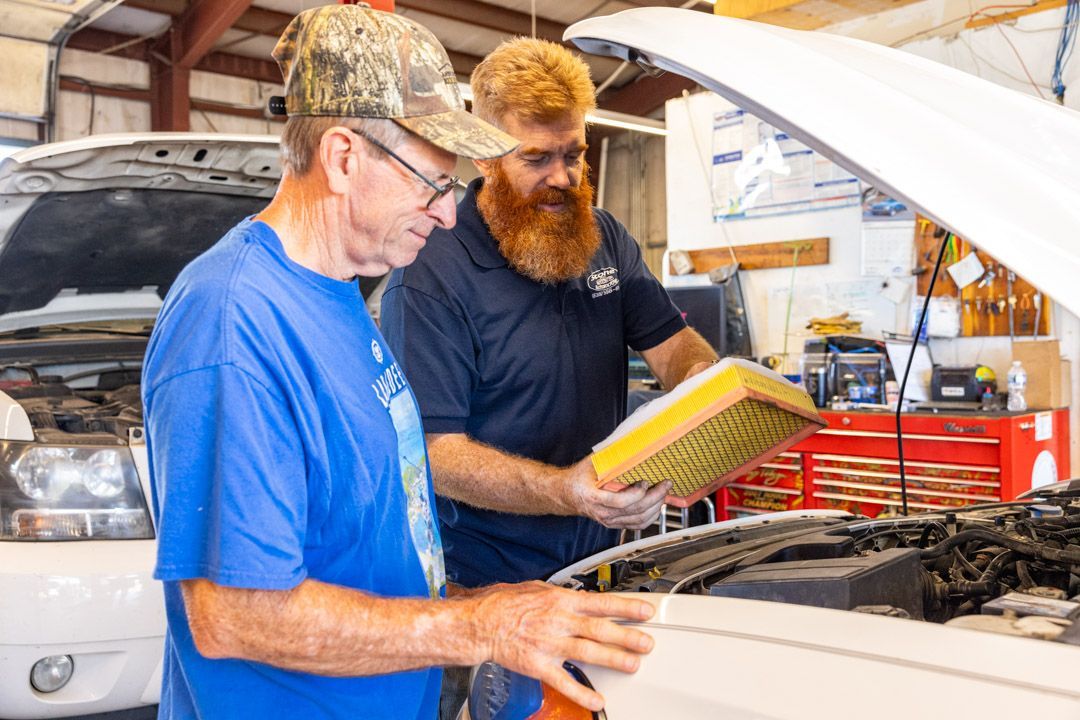 Mechanic showing a car air filter to a customer in a garage. The customer wears a cap and blue shirt.