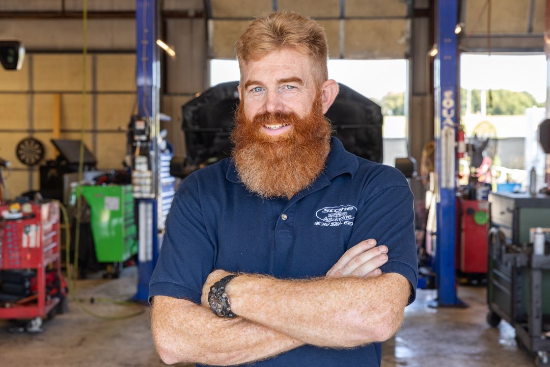 Man with red beard, arms crossed, in a mechanic shop; blue shirt, tools visible.