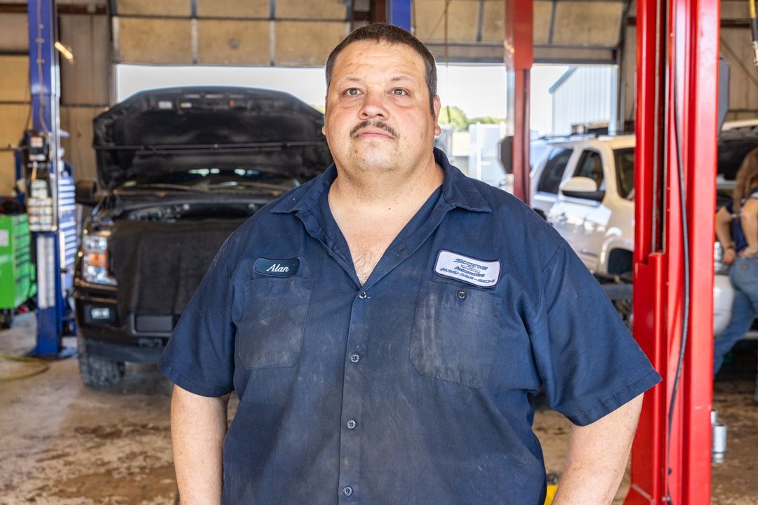 Mechanic in blue work shirt stands in auto shop, smiling, with car behind him.