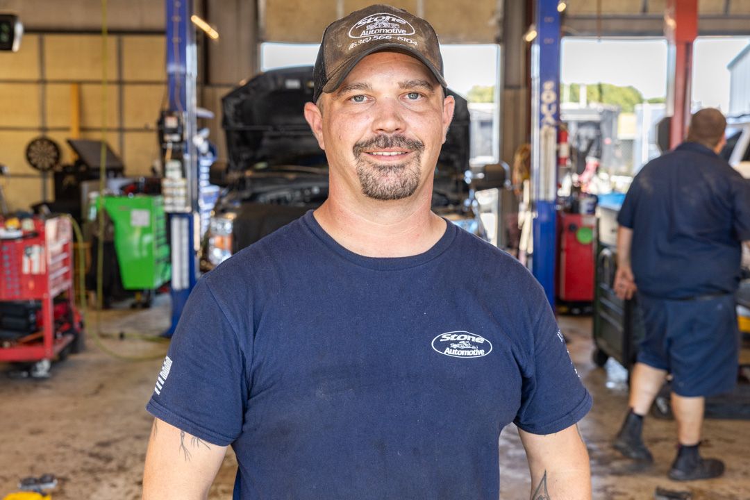 Man in a mechanic shop, wearing a cap and blue shirt, smiling. Another man works in the background.