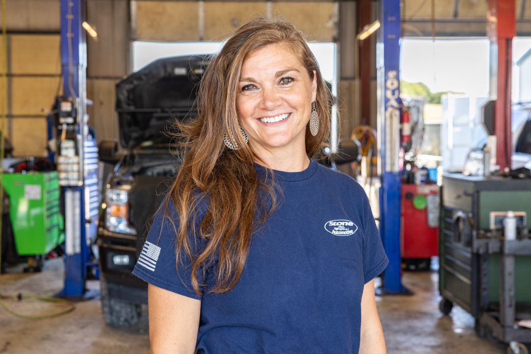 Woman smiling in a car repair shop, wearing a blue shirt with logo, car behind her.