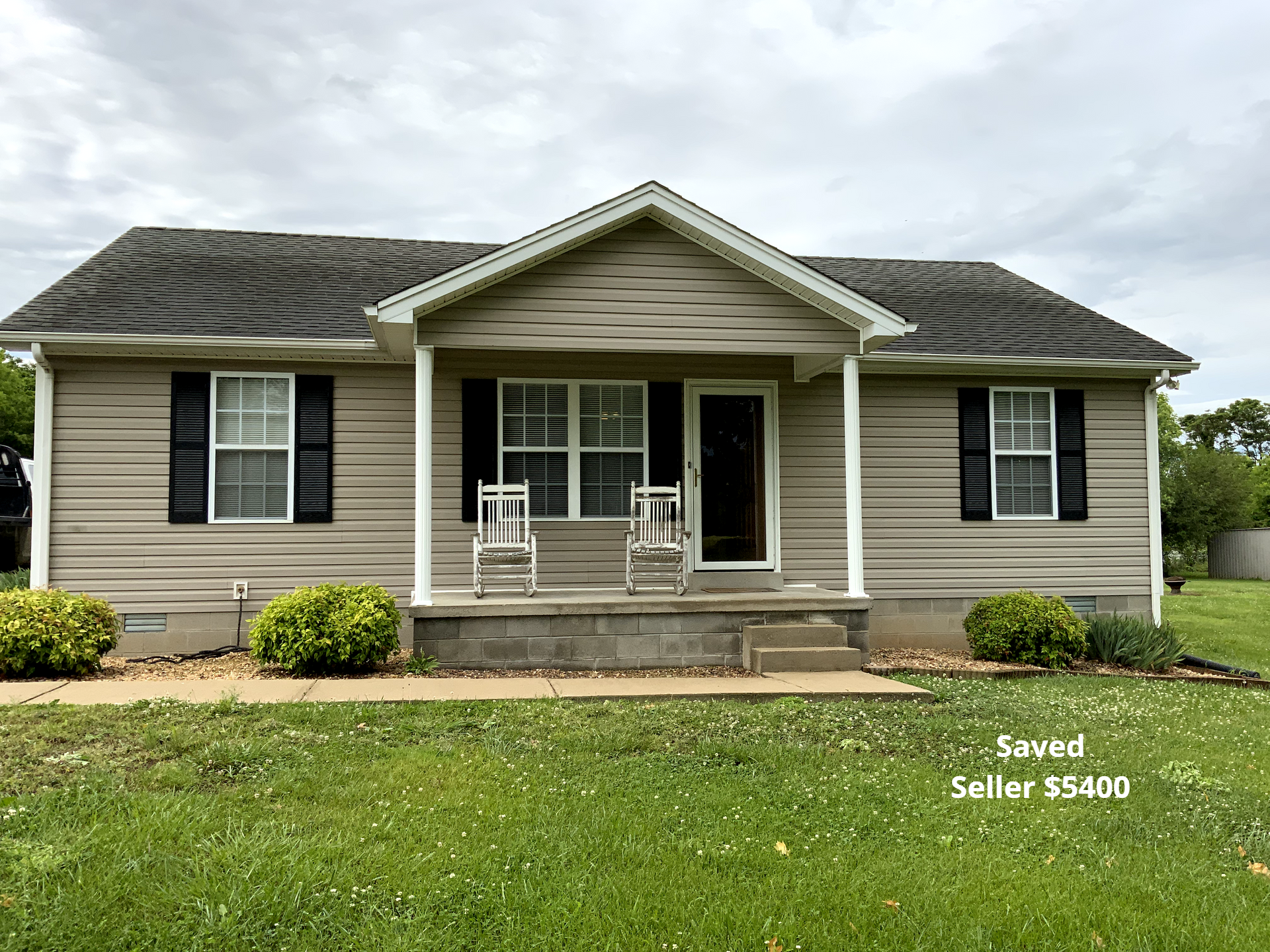 Tan bungalow house with dark shutters, a porch, and green lawn.