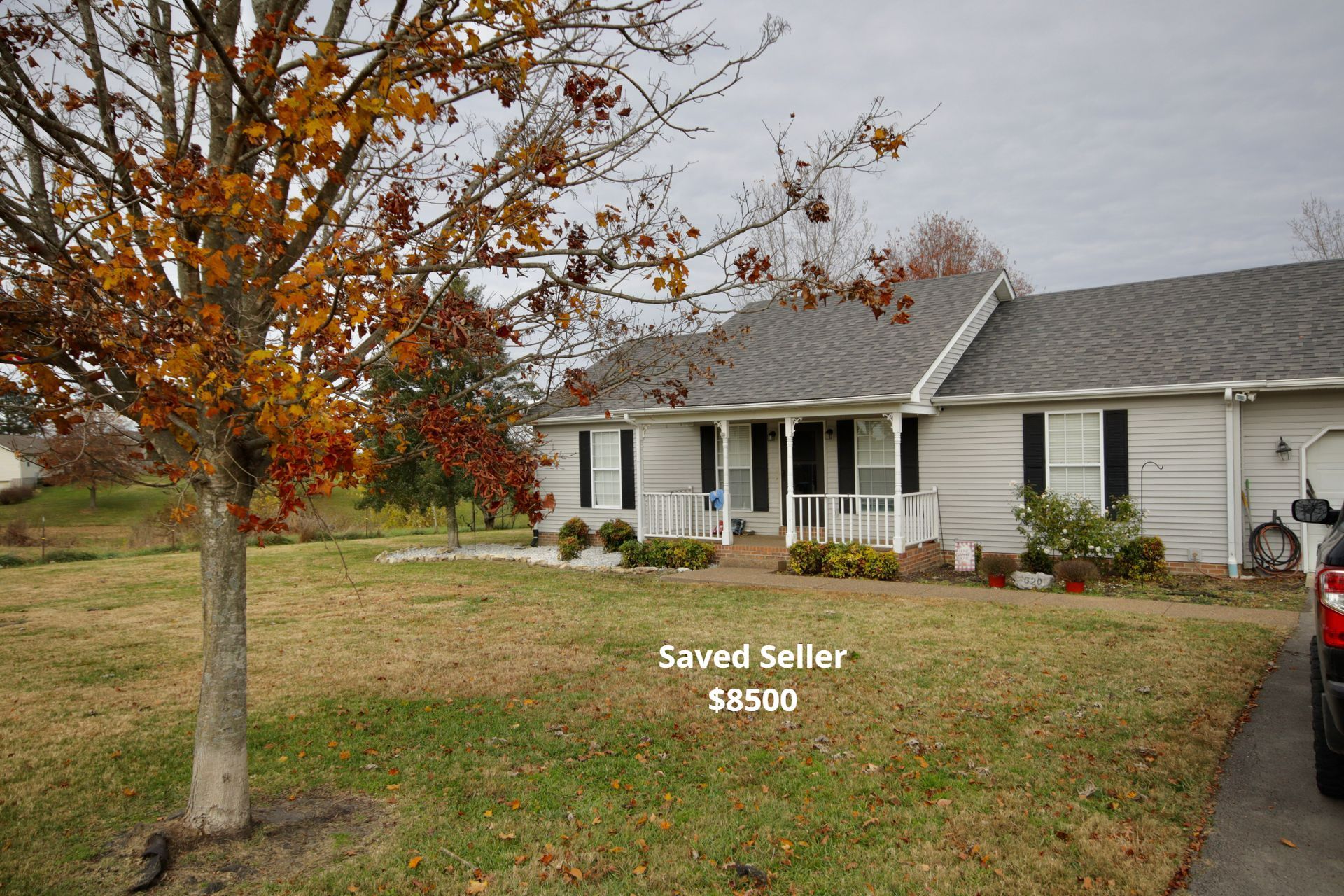 A single-story house with a gray roof and beige siding sits on a grassy lawn, a tree with fall foliage in the foreground.